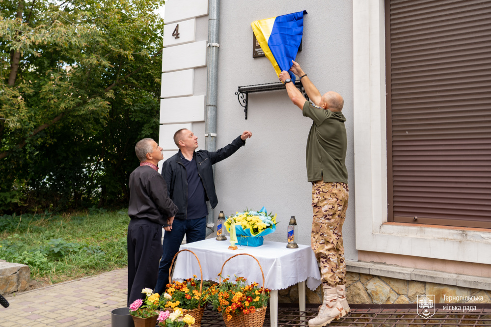 Чоловік знімає синьо-жовте покриття з меморіальної дошки на фасаді будинку. Поруч стоять двоє чоловіків, перед ними стіл із квітами.